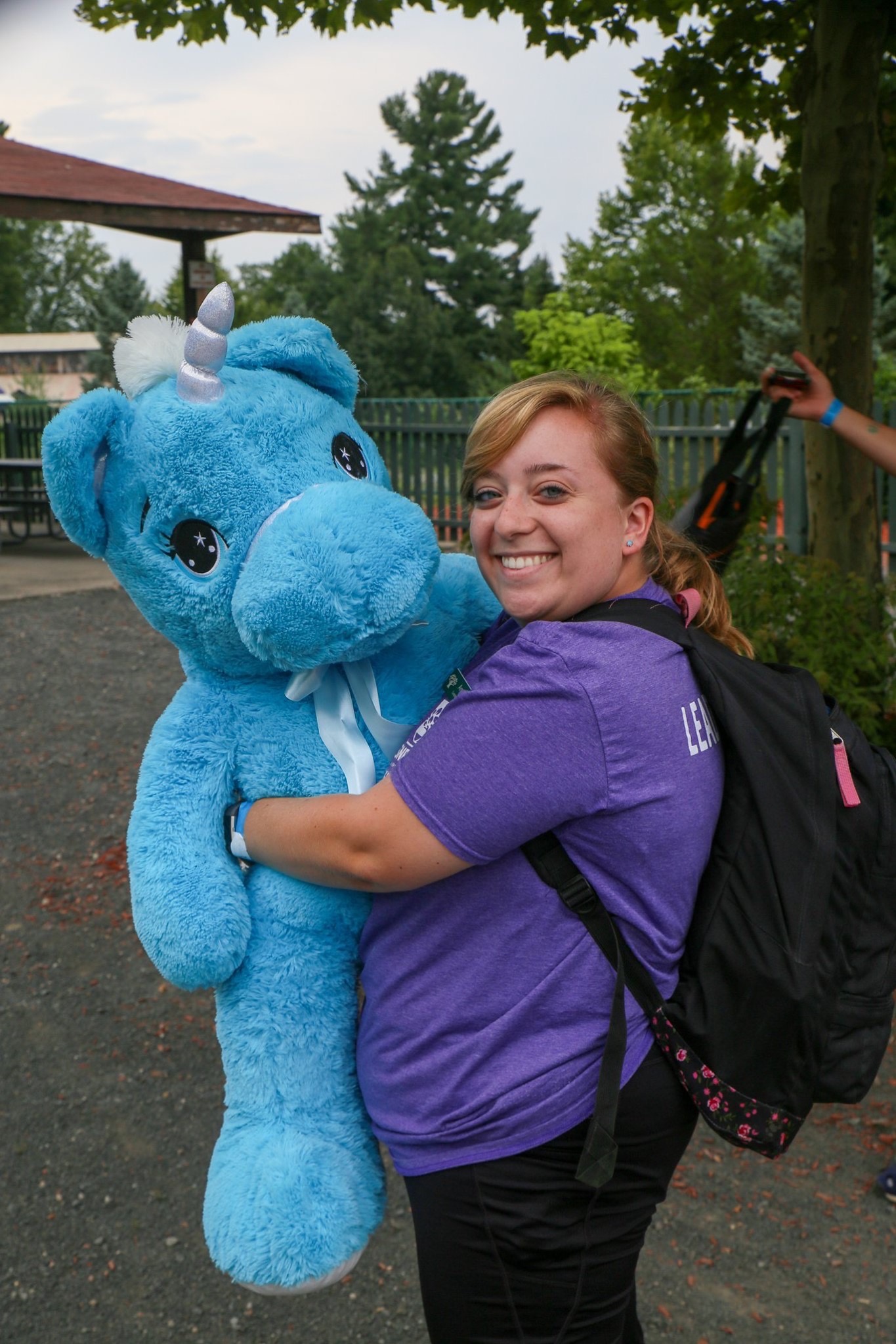 Amanda wearing a purple t-shirt, black pants, and a backpack smiling while hugging an oversized blue stuffed unicorn.