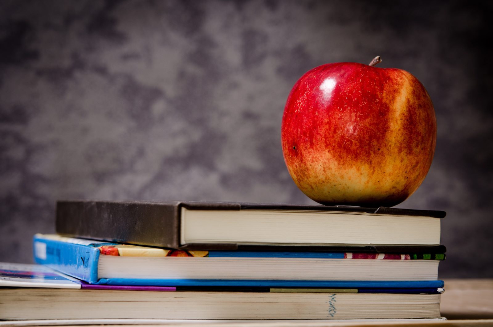 A stack of textbooks with an apple on top and a chalkboard in the background.