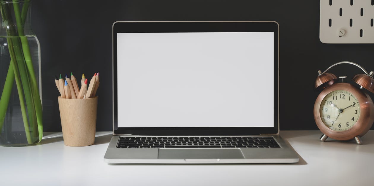 An open laptop on a desk with a jar of colored pencils, a plant, and an alarm clock.