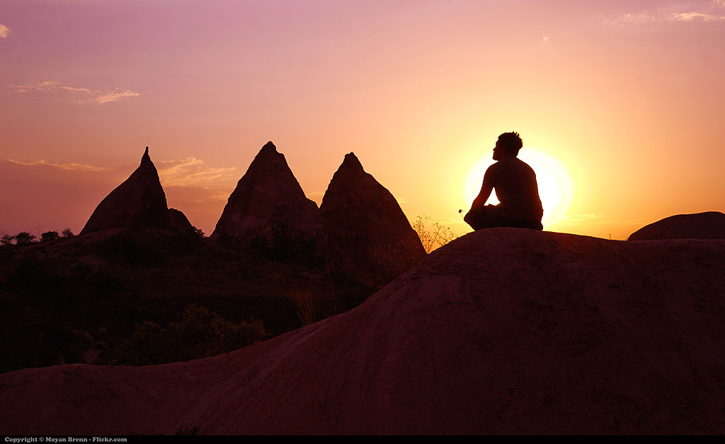 A man sitting on top of a hill looking off into the sunset with mountains in the background