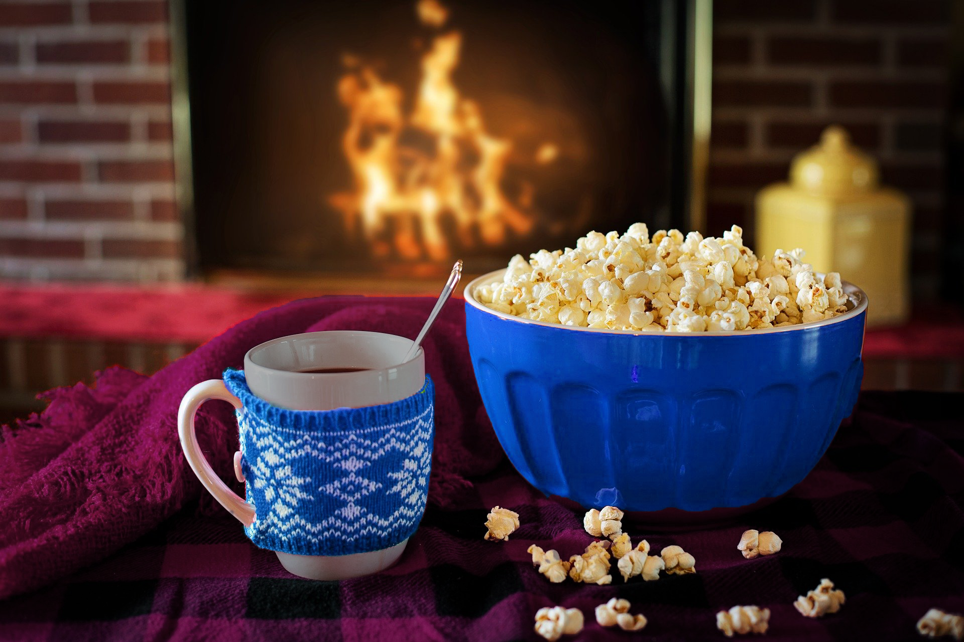 A bowl of popcorn and a mug of hot chocolate on a blanket in front of a fireplace.